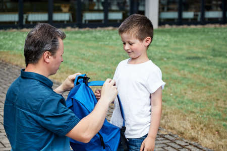 Dad, dad puts a blue backpack on his son. Parents accompany their children to school, first grade. Back to school after summer breakの写真素材