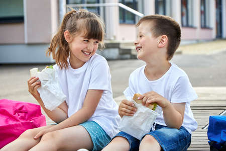 Schoolchildren boy and girl laugh and eat their lunch, snack, breakfast in the school yard. Food for children in educational institutions, kindergartens.の写真素材