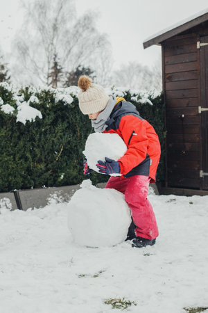 Snow games, entertainment and fun. A cute Caucasian girl sculpts a snowman in the yard. Vertical orientation, authentic peopleの写真素材