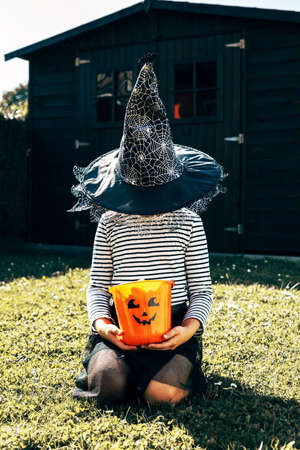 Halloween concept. A girl in a witch costume and a hat that covers her face sits on the grass and holds an orange pumpkin bucket in her hands. Copy space. Vertical photoの写真素材