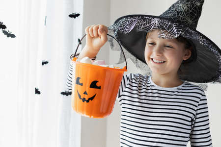Halloween concept. A girl in a witch hat holds an orange pumpkin bucket with sweets and smiles. Kids tricks or treatsの写真素材
