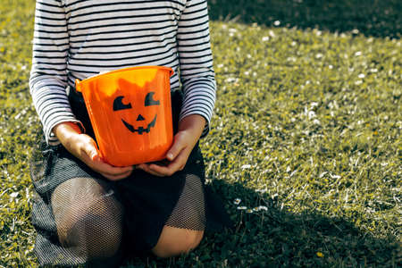 Halloween concept. A girl in a suit sits on the grass and holds an orange pumpkin bucket in her hands. Copy spaceの写真素材