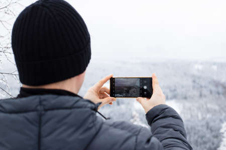 Close-up of a man photographing a winter landscape on the phone. Mobile photography. Winter tourism and hiking in the forest. Copy spaceの写真素材