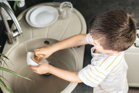 cute boy washes dishes with a wooden brush with natural bristles in the kitchen
