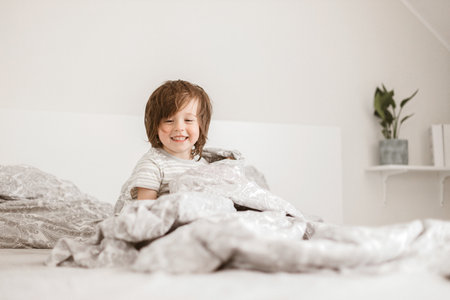 An adorable boy, Toddler, plays on the parents bed. Sits wrapped in a blanket and laughs. Family fun