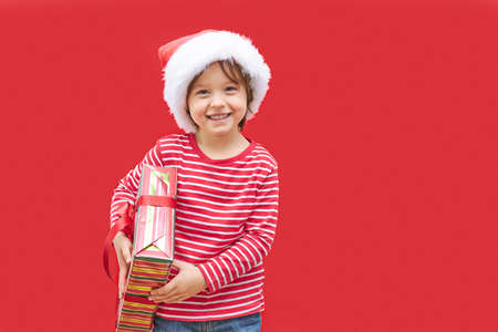 A charming boy in a santa claus hat and a gift in his hands is standing on a red background and smiling. Shopping for Christmas and New Years. Congratulationsの写真素材