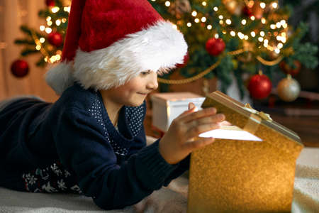 A boy in a santa hat opens a gift box next to a garlanded Christmas tree. Gifts for children. Dreams Come Trueの写真素材
