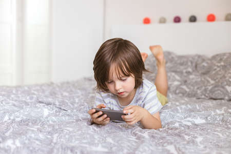 A cute boy, toddler, lies on the bed in the bedroom and plays with the phone, smartphone. Problems...