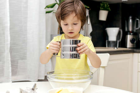Little cute boy adds flour to the dough. Children cook in the kitchen. Little chefsの写真素材