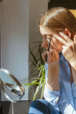Young beautiful girl applies makeup on her face next to the window. Reflection in the mirror, personal and skin care. Daily Makeupの写真素材