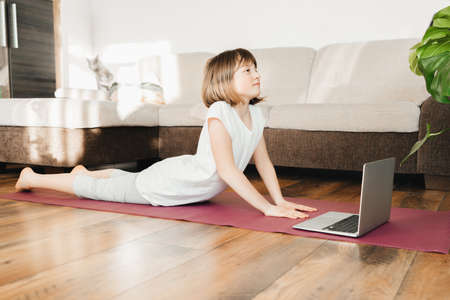 Cute girl goes in for sports at home. Child learns yoga at home online. Childrens sports and...