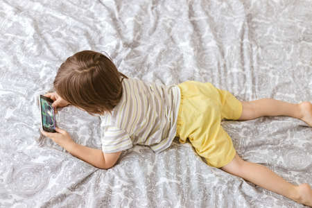 A cute boy, toddler, lies on the bed in the bedroom and plays with the phone, smartphone. Problems of children and gadgets. Video games and internetの写真素材