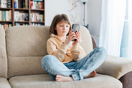 Schoolgirl is sitting on the sofa and playing mobile phone or chatting with friends. Children use...