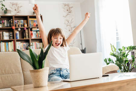 Girl schoolgirl at home in front of a laptop rejoices at success. Online learning, distance education and coursesの写真素材