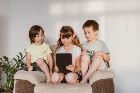 Three children sit on an armchair at home and use a digital tablet. Children and gadgets. Online learning and communication. Use of technology in the modern world.の写真素材