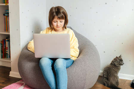 Children and gadgets. A girl is sitting in a chair in her room with a laptop on her lap and chatting with friends or watching an educational webinar.の写真素材