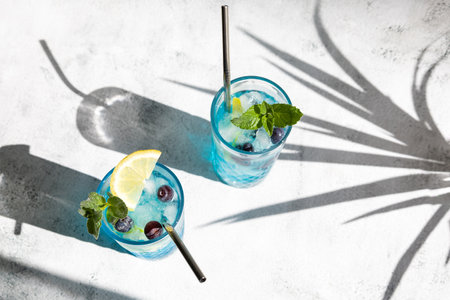 Curacao blue cocktail with fresh blueberries and lemon wedges, garnished with mint with trendy shadows. Summer refreshing soft drink. View from aboveの写真素材