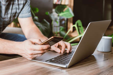 Close-up of a phone in the hands of a man next to a laptop at home. Work online. Online shopping and payment using gadgets.の写真素材