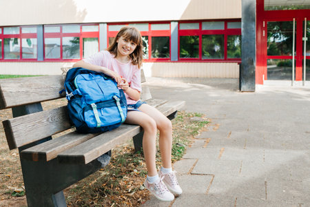 Back to school. A schoolgirl teenager sits on a bench next to a schoolbag in front of the school, smiling and looking at the camera.の写真素材