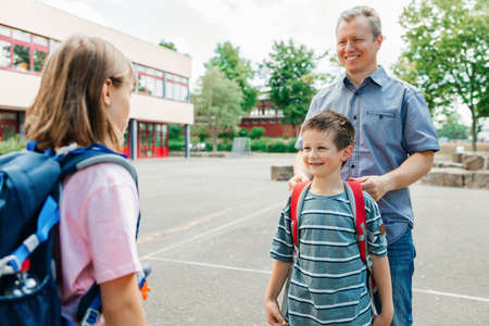 Back to school. A happy dad escorts his children, a boy and a girl, to school, helps his son put on a backpack. Parental care for childrenの写真素材
