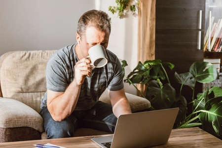 A young pensive man sits at home in an armchair and works on a laptop. Man drinking coffee and...
