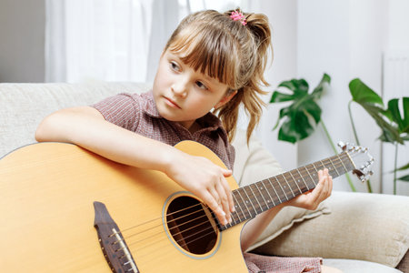 A cute little girl is sitting on the couch at home and learning to play the acoustic guitar. Hobbies...