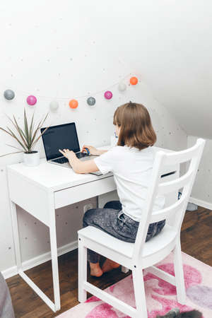 A teenage girl is sitting at a table in her room and working on a laptop, watching webinars or chatting with friends. The role of gadgets in the life of modern children.の写真素材