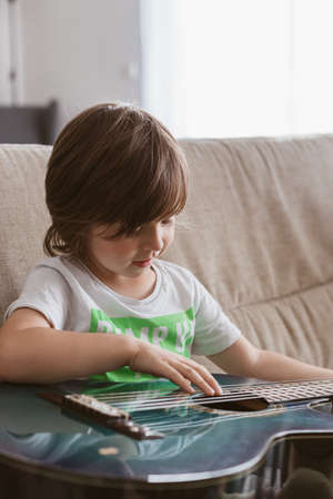 Funny little boy is learning to play the acoustic guitar at home, sitting on the couch and laughing.の写真素材