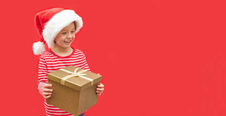 A little boy in a santa claus hat and a large gift box in his hands stands on a red background. Bannerの写真素材