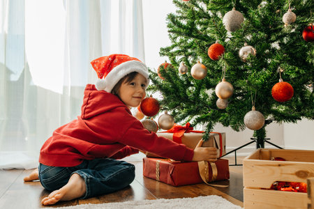 Charming boy in a santa hat takes out Christmas gifts from under the New Year tree and laughsの写真素材