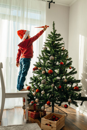 A boy in a santa claus hat decorates the New Year tree at home, puts a Christmas toy on the top of the tree. Children are preparing to celebrate Christmas and New Yearの写真素材
