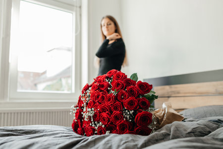 A huge beautiful bouquet of scarlet roses on a bed in a bedroom or hotel and a beautiful girl in the background by the window. Flowers for the holidaysの写真素材