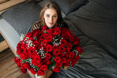 A beautiful girl is sitting on a bed in the bedroom with a huge bouquet of scarlet roses. Flower delivery to women on holidays and memorable datesの写真素材