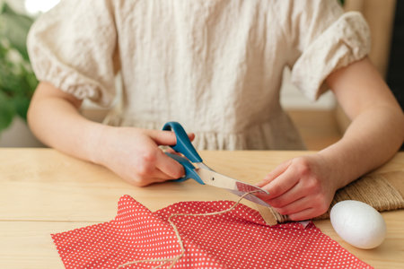 A girl at home makes Easter egg decorations in the shape of a rabbit from fabric. The child cuts the...