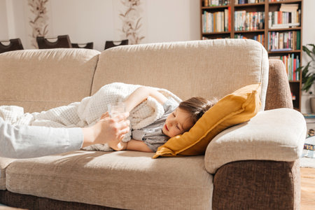 A little sick boy lies on the couch and his mother holds out a glass of water to the child. The period of epidemics of influenza and seasonal diseases.の写真素材