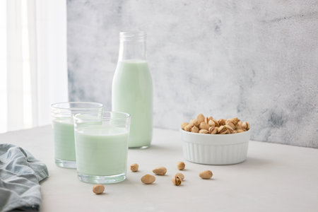 A glass of plant-based nut milk is on the table next to a bottle of pistachio milk and pistachios....