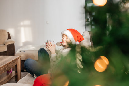 A teenage girl drinks hot chocolate or cocoa while sitting on the sofa in the living room next to the Christmas tree. Enjoyment and relaxation while waiting for New Year and Christmas.の写真素材