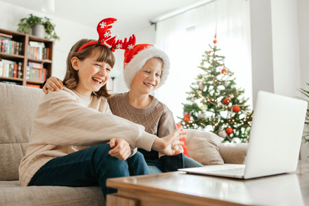 Children teenagers boy and girl sitting on the sofa in the apartment in Santa hats and communicating with family via laptop. Christmas greetings onlineの写真素材