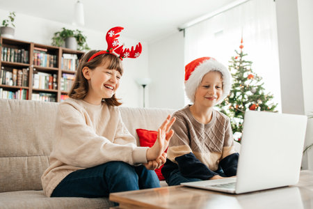 Children teenagers boy and girl sitting on the sofa in the apartment in Santa hats and communicating with family via laptop. Christmas greetings onlineの写真素材