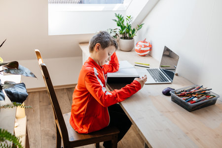 A boy reading a book sitting at a table in a room. The child is engrossed in reading or doing...