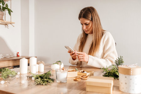 A woman takes a photo of Christmas gifts and decorations on a table with her smartphone, a cozy home atmosphere, and preparations for the holiday.の写真素材