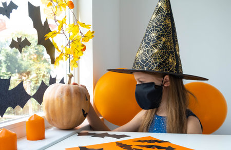 Little sad girl in black face mask and witch costume looking at the pumpkin in decorated room. Kid in black hat preparing to celebrate Halloween at home. Protecting from COVID-19.の写真素材