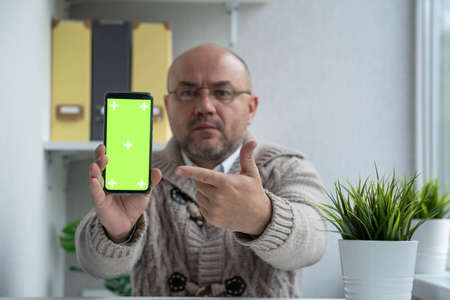 Serious man with glasses pointing finger on the phone with a green screen. Chroma key green screen...