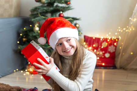 A blond haired little girl holding Christmas gift near the Christmas Tree. Trying to understand what is in the package, shakes present next to her ear. Listens to guess the contents with a smile.の写真素材