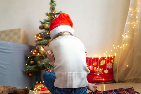 Sad little girl wearing Santa hat sitting with back turned on the background of decorated Christmas tree disappointed with gift. Cute little child feeling unhappy with bad holiday present.の写真素材