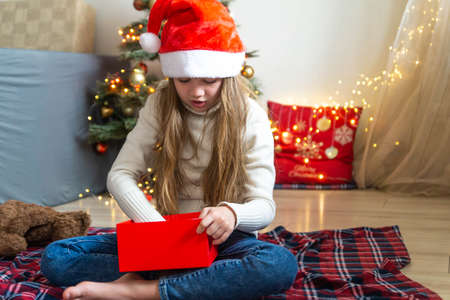 Cute little girl in Santa hat preparing Christmas gift on the background of decorated tree at home. DIY present, holiday concept. New Year celebration.の写真素材