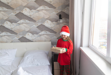 Little child girl in red hat happy with gift from Santa on Christmas morning. Kid with present at home in parents bedroom. Magic holidays, happy childhood. Happy new year.の写真素材