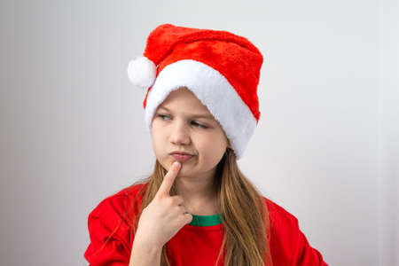 Pensive little girl in Christmas red hat on white background. Serious child thinking, looking sideways with doubtful and skeptical expression. New Year gift shopping, making wishes for Santa Claus.の写真素材