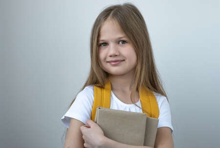 Portrait of pretty cute smiling school girl in white t-short with yellow bag and books on gray background. Elementary school, education concept.の写真素材