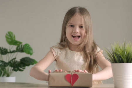 Little girl smiling giving handmade stylish simple gift box in craft paper at home with small red heart. Happy child with present for Valentines Day, mothers or fathers day. DIY, zero waste concept.の写真素材